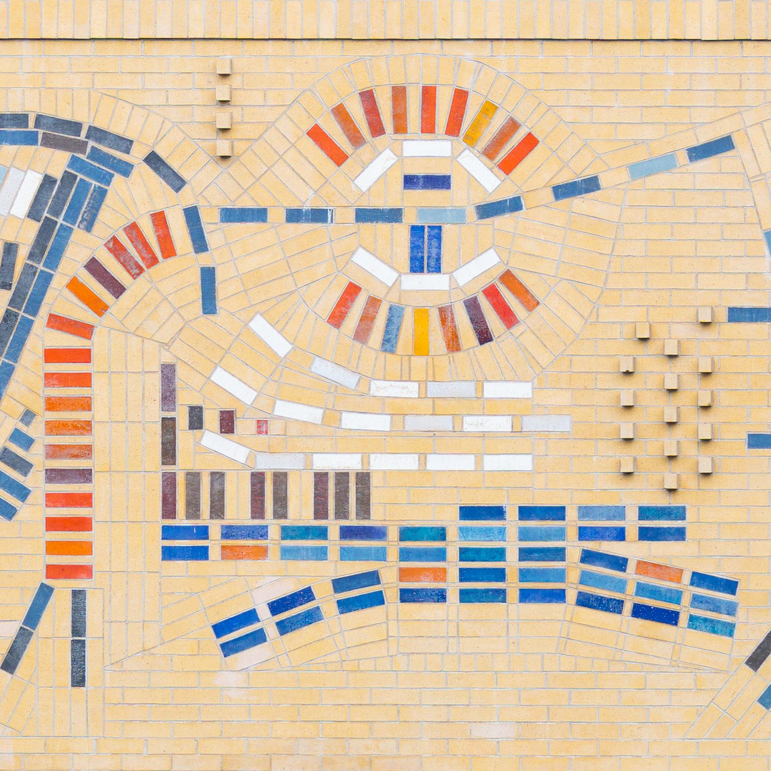 Gallery 4 - Close up of a large, colorful abstract mosaic tile mural on the beige brick wall of a building at Contra Costa College. The artwork features stylized, interconnected symbols in blue, red, orange, and white, suggesting themes of science, nature, and evolution. A grassy lawn and a road are in the foreground, with trees and hills in the background.