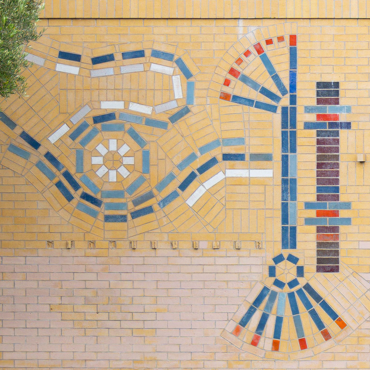 Gallery 7 - A large, colorful abstract mosaic tile mural on the beige brick wall of a building at Contra Costa College. The artwork features stylized, interconnected symbols in blue, red, orange, and white, suggesting themes of science, nature, and evolution. A grassy lawn and a road are in the foreground, with trees and hills in the background.