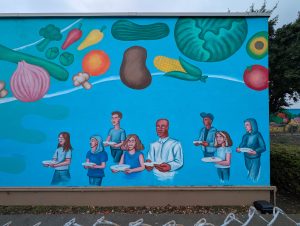 Mural on a wall with a blue background and people holding empty plates, and there are floating vegetables at the top.