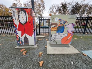 A grandfather clock covers one Utility Box, surrounded by red swirls, music notes, and a row of colorful flowers. On the box next to it, a blue dog plays a piano while a pink dog in glasses plays a cello above a line of music.