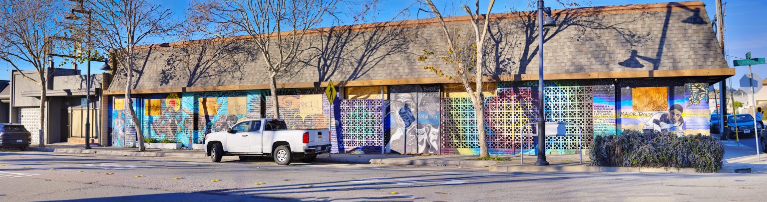 Gallery 1 - A wide, multi-panel mural wrapping around a building featuring colorful figures, portraits, and geometric patterns. Sections include a portrait of Bayard Rustin, a cyclist, a woman flexing, and vibrant rainbows and sunflowers.