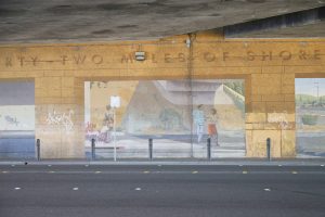 A mural on a concrete underpass wall showing a vintage streetcar and passengers from 1908 appearing as a reflection in a puddle. Figures in modern clothing, including a person pushing a shopping cart, stand near figures in historic dress.