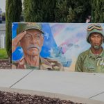 Gallery 1 - A wide outdoor mural on a low concrete wall depicting six military service members in various uniforms, including a veteran in a saluting pose. The center includes the message 