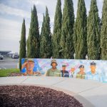 Gallery 2 - A wide outdoor mural on a low concrete wall depicting six military service members in various uniforms, including a veteran in a saluting pose. The center includes the message 