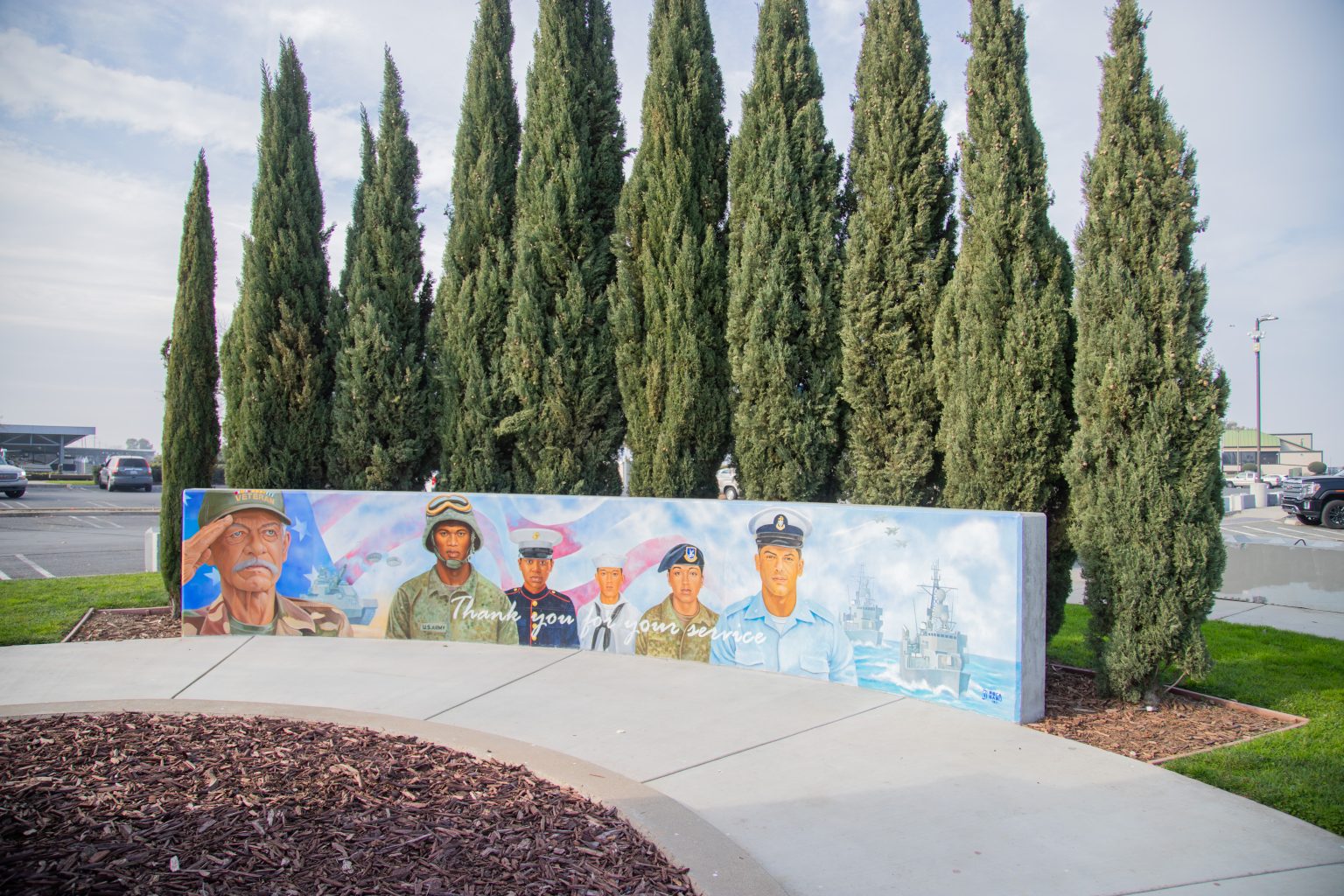 Gallery 2 - A wide outdoor mural on a low concrete wall depicting six military service members in various uniforms, including a veteran in a saluting pose. The center includes the message 