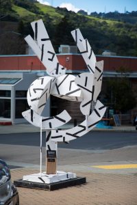 A white abstract steel sculpture with black linear markings stands on a sidewalk. The piece features curved and angular interlocking plates that create a sense of movement and negative space.