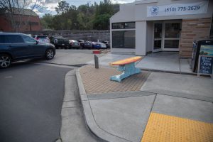 A functional bench shaped like a surfboard, covered in colorful ceramic mosaic tiles. The top is red and yellow with jagged patterns, while the base is composed of various shades of blue and turquoise tiles.