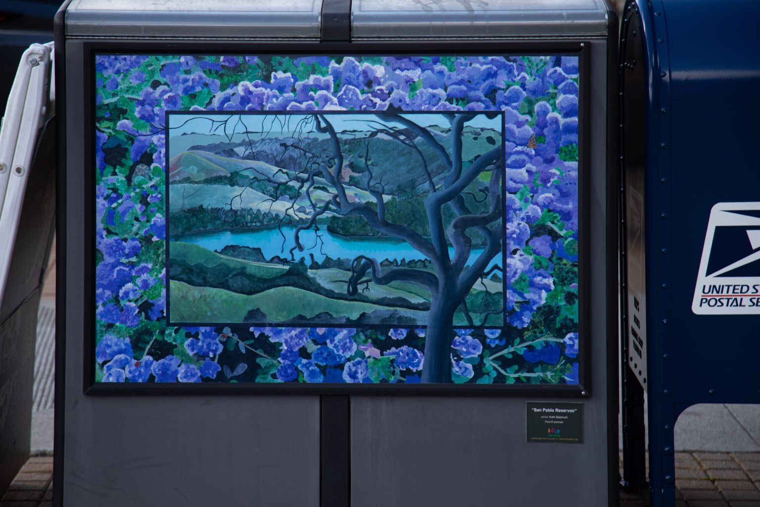 Gallery 1 - A utility box features a landscape painting of the San Pablo Reservoir viewed through the branches of a dark, twisting tree. The central image is framed by a dense border of vibrant purple hydrangeas, and a small plaque encourages viewers to find six hidden animals.