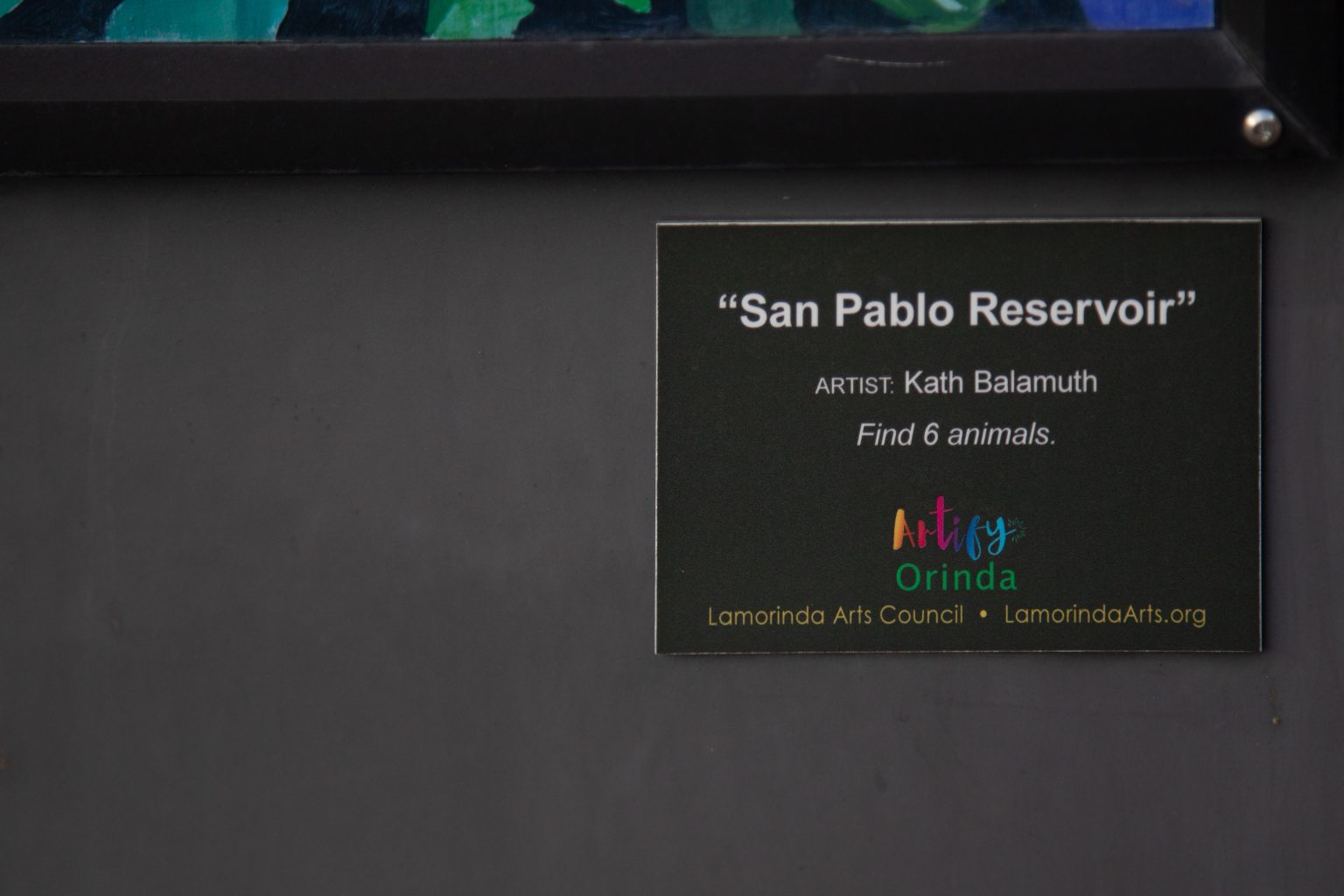 Gallery 2 - A plaque of utility box features a landscape painting of the San Pablo Reservoir viewed through the branches of a dark, twisting tree. The central image is framed by a dense border of vibrant purple hydrangeas, and a small plaque encourages viewers to find six hidden animals.