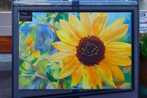 A vibrant watercolor painting of a sunflower with bright yellow petals and a textured brown center. The background features loose brushstrokes of green leaves and a blue sky.