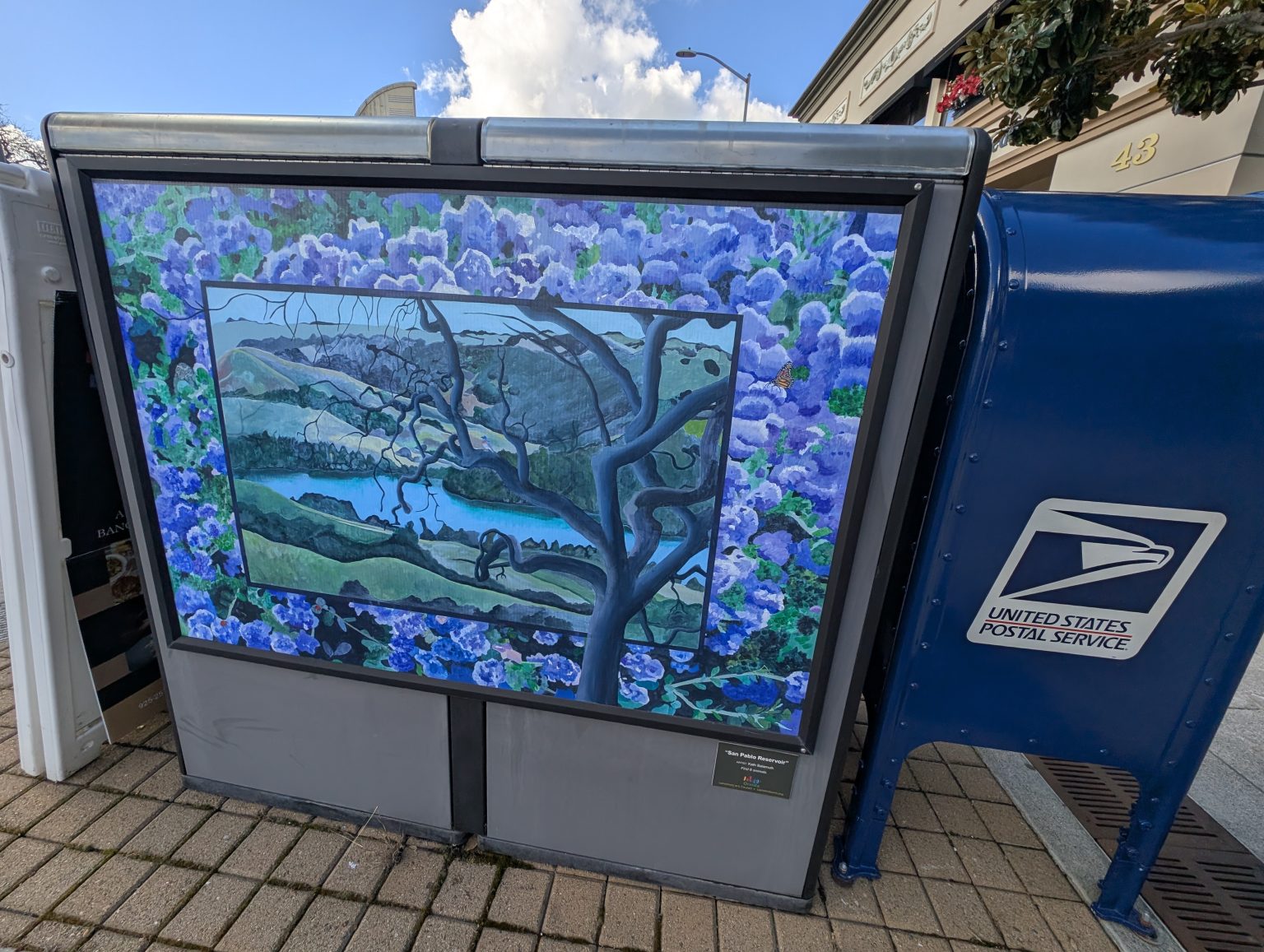 Gallery 3 - A utility box features a landscape painting of the San Pablo Reservoir viewed through the branches of a dark, twisting tree. The central image is framed by a dense border of vibrant purple hydrangeas, and a small plaque encourages viewers to find six hidden animals.
