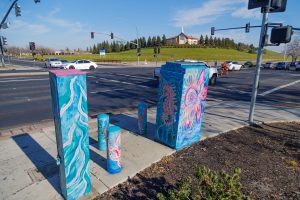 A utility box and three surrounding bollards painted with a vibrant underwater scene. The central box features a large pink seahorse, while the bollards and base show smaller fish and sea plants in shades of blue and teal.
