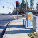 Gallery 7 - Painted utility box at a street corner with large orange flowers on a light blue background. The artwork brightens a traffic signal cabinet beside Sagebrush Drive in Antioch.
