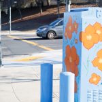 Gallery 6 - Painted utility box at a street corner with large orange flowers on a light blue background. The artwork brightens a traffic signal cabinet beside Sagebrush Drive in Antioch.