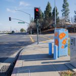 Gallery 5 - Painted utility box at a street corner with large orange flowers on a light blue background. The artwork brightens a traffic signal cabinet beside Sagebrush Drive in Antioch.