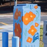 Gallery 4 - Painted utility box at a street corner with large orange flowers on a light blue background. The artwork brightens a traffic signal cabinet beside Sagebrush Drive in Antioch.