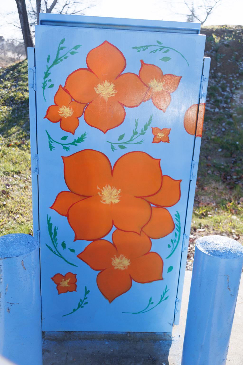 Gallery 3 - Painted utility box at a street corner with large orange flowers on a light blue background. The artwork brightens a traffic signal cabinet beside Sagebrush Drive in Antioch.