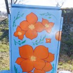 Gallery 3 - Painted utility box at a street corner with large orange flowers on a light blue background. The artwork brightens a traffic signal cabinet beside Sagebrush Drive in Antioch.