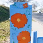 Gallery 2 - Painted utility box at a street corner with large orange flowers on a light blue background. The artwork brightens a traffic signal cabinet beside Sagebrush Drive in Antioch.
