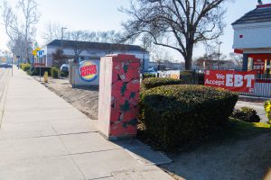 A utility box on a sidewalk is painted with a dense pattern of large red strawberries. The strawberries have white seeds and green leafy tops, covering the sides of the metal box.