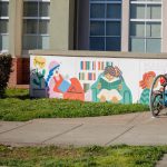 Gallery 1 - A colorful outdoor mural on a low concrete wall in front of a school building. It features stylized figures of children and adults reading, playing music, and participating in a 
