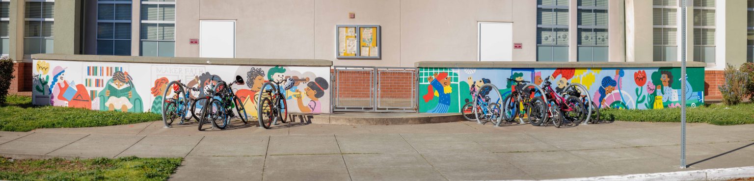 Gallery 1 - A colorful outdoor mural on a low concrete wall in front of a school building. It features stylized figures of children and adults reading, playing music, and participating in a 