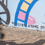 Gallery 2 - A colorful outdoor mural on a low concrete wall in front of a school building. It features stylized figures of children and adults reading, playing music, and participating in a 