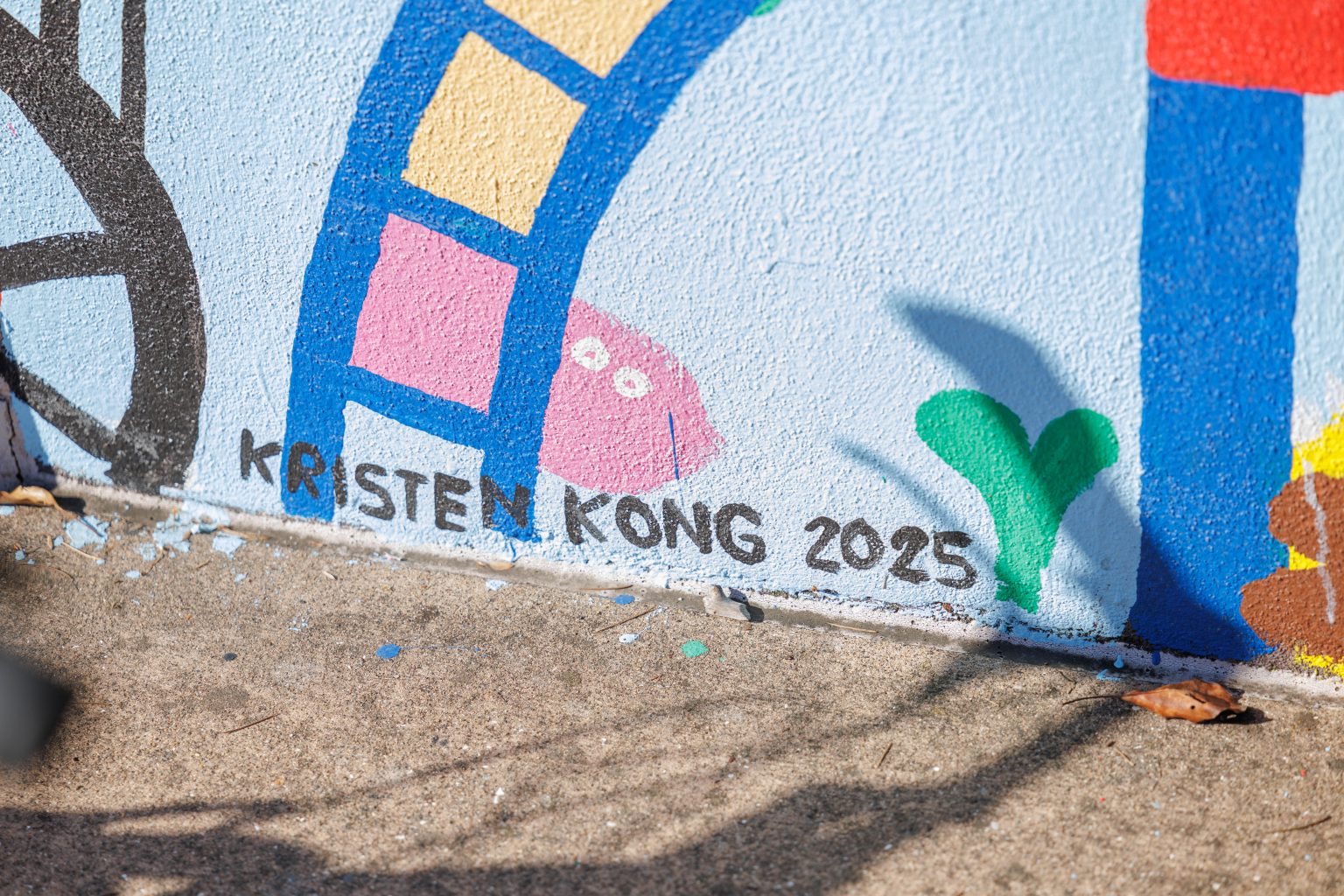 Gallery 2 - A colorful outdoor mural on a low concrete wall in front of a school building. It features stylized figures of children and adults reading, playing music, and participating in a 