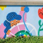 Gallery 3 - A colorful outdoor mural on a low concrete wall in front of a school building. It features stylized figures of children and adults reading, playing music, and participating in a 