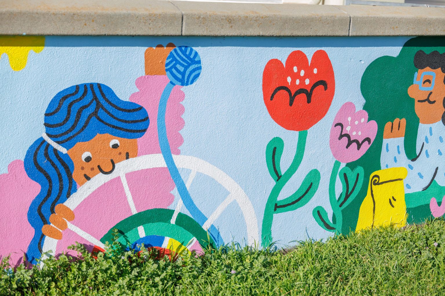 Gallery 3 - A colorful outdoor mural on a low concrete wall in front of a school building. It features stylized figures of children and adults reading, playing music, and participating in a 