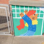 Gallery 4 - A colorful outdoor mural on a low concrete wall in front of a school building. It features stylized figures of children and adults reading, playing music, and participating in a 