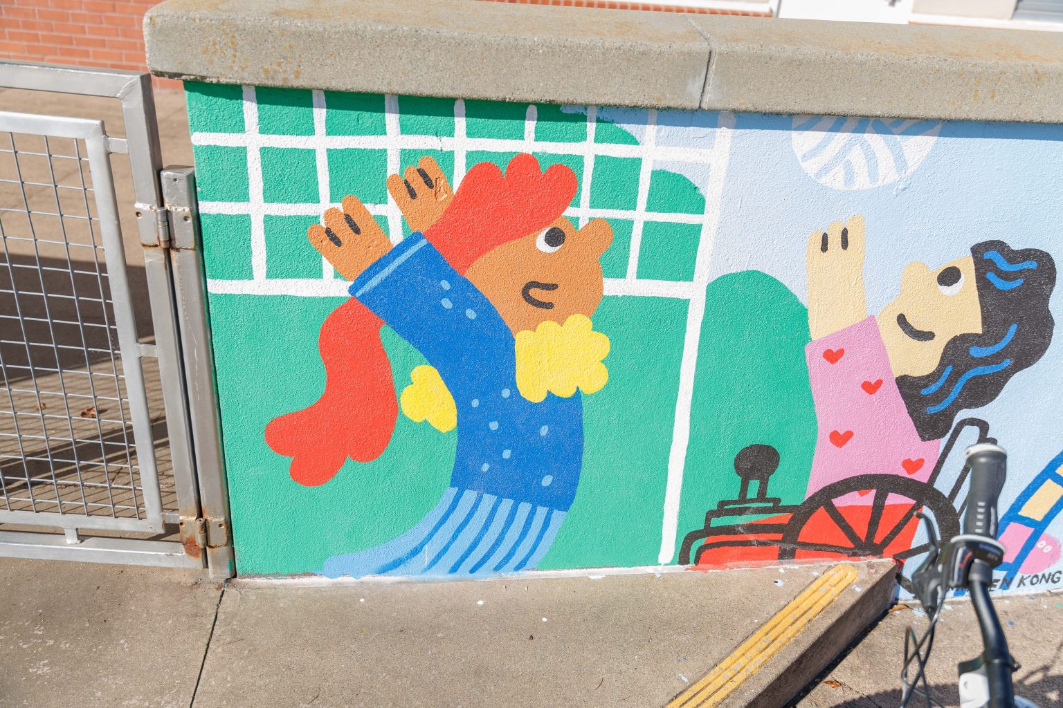 Gallery 4 - A colorful outdoor mural on a low concrete wall in front of a school building. It features stylized figures of children and adults reading, playing music, and participating in a 