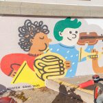 Gallery 5 - A colorful outdoor mural on a low concrete wall in front of a school building. It features stylized figures of children and adults reading, playing music, and participating in a 