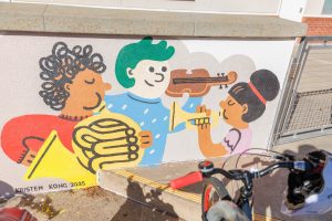 A colorful outdoor mural on a low concrete wall in front of a school building. It features stylized figures of children and adults reading, playing music, and participating in a "Maker Fair."