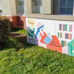 Gallery 6 - A colorful outdoor mural on a low concrete wall in front of a school building. It features stylized figures of children and adults reading, playing music, and participating in a 