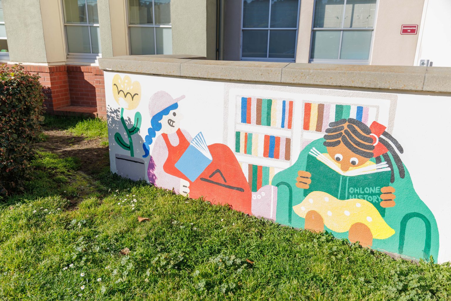 Gallery 6 - A colorful outdoor mural on a low concrete wall in front of a school building. It features stylized figures of children and adults reading, playing music, and participating in a 