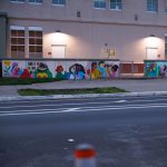 Gallery 7 - A colorful outdoor mural on a low concrete wall in front of a school building. It features stylized figures of children and adults reading, playing music, and participating in a 