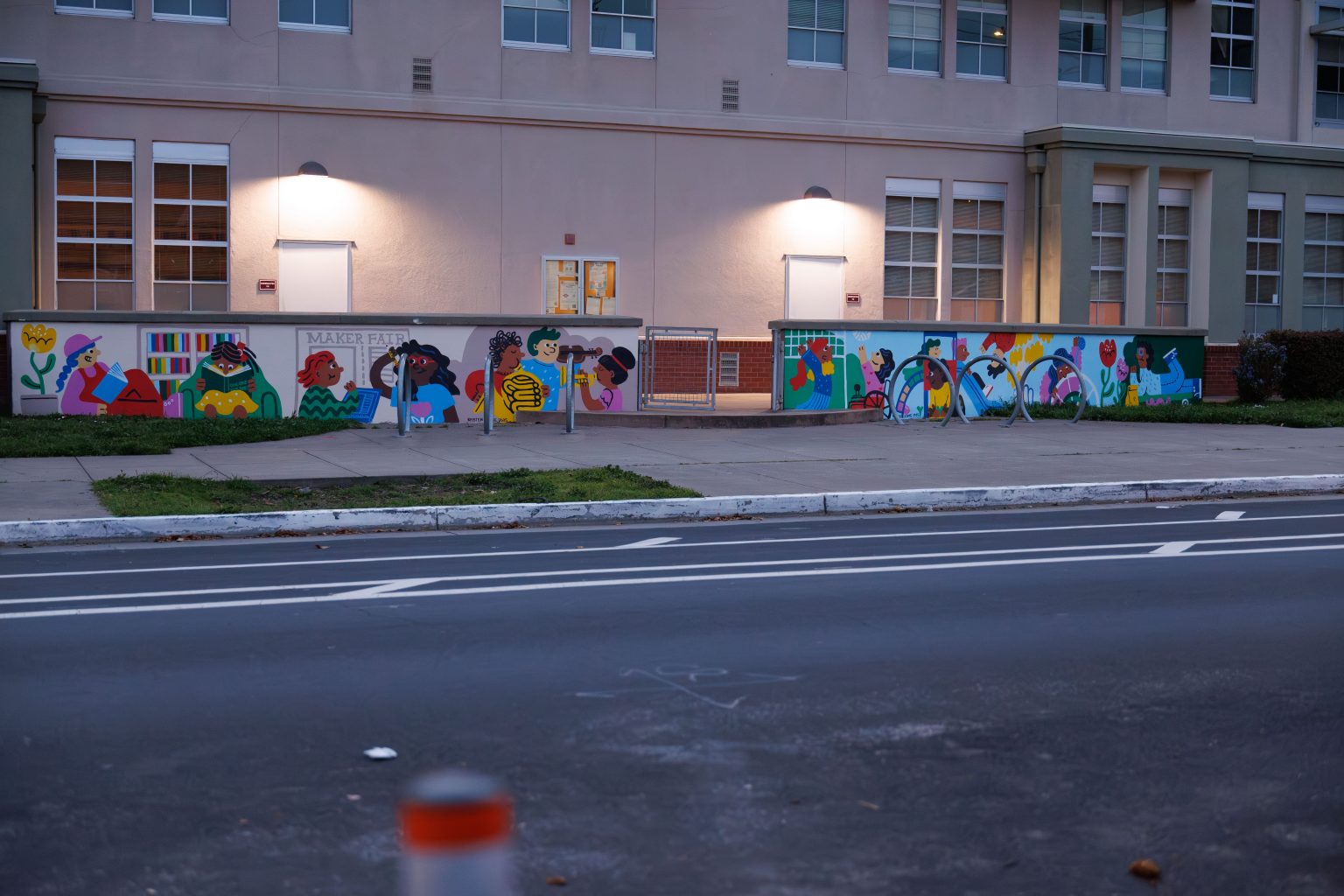 Gallery 7 - A colorful outdoor mural on a low concrete wall in front of a school building. It features stylized figures of children and adults reading, playing music, and participating in a 