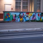 Gallery 8 - A colorful outdoor mural on a low concrete wall in front of a school building. It features stylized figures of children and adults reading, playing music, and participating in a 