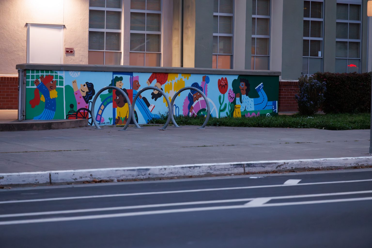 Gallery 8 - A colorful outdoor mural on a low concrete wall in front of a school building. It features stylized figures of children and adults reading, playing music, and participating in a 