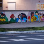Gallery 9 - A colorful outdoor mural on a low concrete wall in front of a school building. It features stylized figures of children and adults reading, playing music, and participating in a 