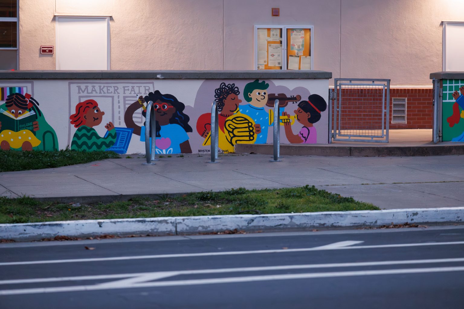 Gallery 9 - A colorful outdoor mural on a low concrete wall in front of a school building. It features stylized figures of children and adults reading, playing music, and participating in a 