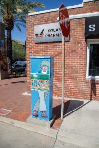 A painted utility box shows a person in a bucket hat holding a sign that says “PITTSBURG,” with a green landscape painted behind them.