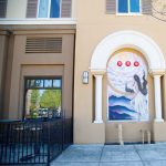 Gallery 2 - A large-scale mural on the side of a commercial building featuring bold, colorful portraiture blended with geometric shapes and flora. The composition uses a warm palette of oranges and teals to highlight the intersection of the city's industrial heritage and its community growth.