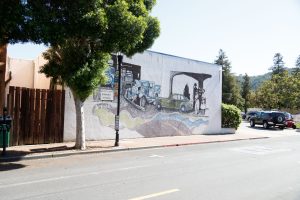 A wide outdoor mural on a white building wall featuring a vintage green car and historical figures in front of a service station. A winding, ribbon-like path along the bottom contains numerous handwritten names of artists and contributors.