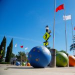 Gallery 3 - A row of seven tall silver flagpoles with red and white flags stands along a school walkway. At the base of each pole is a large concrete sphere painted in vibrant blue, green, or orange, decorated with golden handprints.