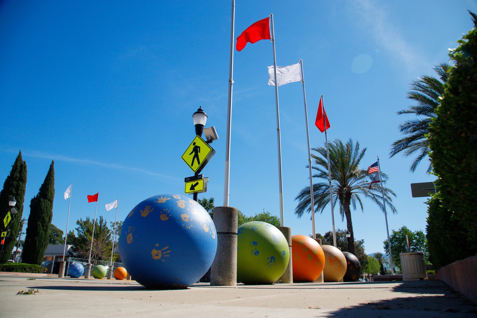 Gallery 3 - A row of seven tall silver flagpoles with red and white flags stands along a school walkway. At the base of each pole is a large concrete sphere painted in vibrant blue, green, or orange, decorated with golden handprints.