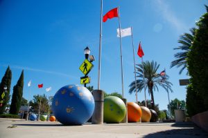 A row of seven tall silver flagpoles with red and white flags stands along a school walkway. At the base of each pole is a large concrete sphere painted in vibrant blue, green, or orange, decorated with golden handprints.