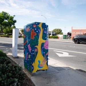 A painted utility box showing a yellow tree, pink flowers, and birds on a blue background with white dots. The box stands on a concrete sidewalk next to a street.