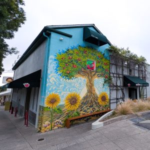 A colorful outdoor mural painted on a light blue building wall beneath a dark green awning marked "EST 1969". The mural depicts a large, vibrant apple tree with deep, twisting roots, flanked by three large yellow sunflowers and small mushrooms at the base, with a red and green neon apple sign glowing in the center of the tree branch canopy.